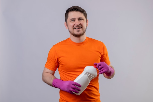 Young man wearing orange t-shirt and rubber gloves holding cleaning supplies smiling positive and happy standing over white wall