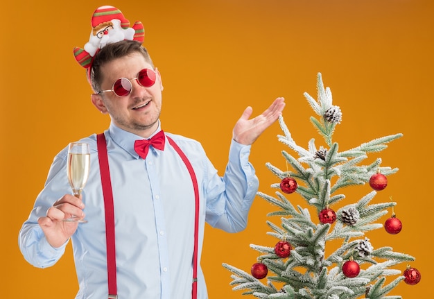 Free Photo young man wearing suspenders bow tie in rim with santa and red glasses standing next to christmas tree holding glass of champagne happy and cheerful over orange background