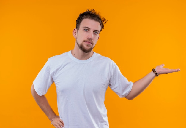 Free photo young man wearing white t-shirt points to side put his hand on hip on isolated orange wall