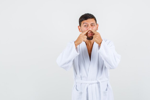 Young man in white bathrobe squeezing acne on his nose, front view.