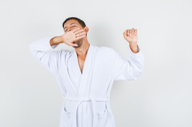 Young man in white bathrobe yawning and stretching and looking relaxed , front view.