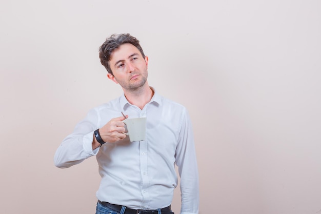 Young man in white shirt, jeans holding cup of drink and looking pensive