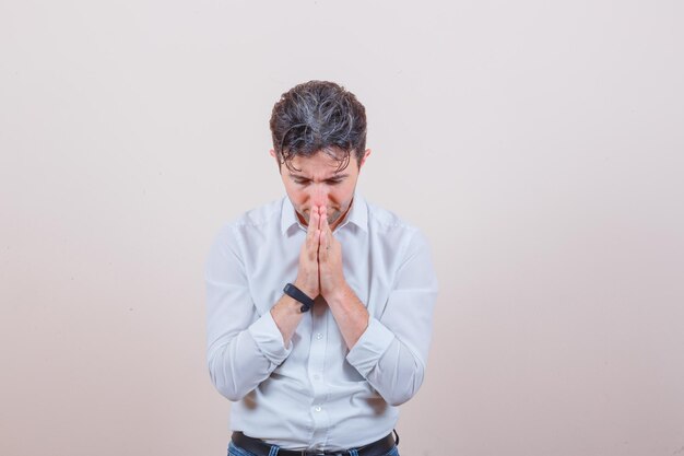 Young man in white shirt, jeans holding hands in praying gesture and looking hopeful