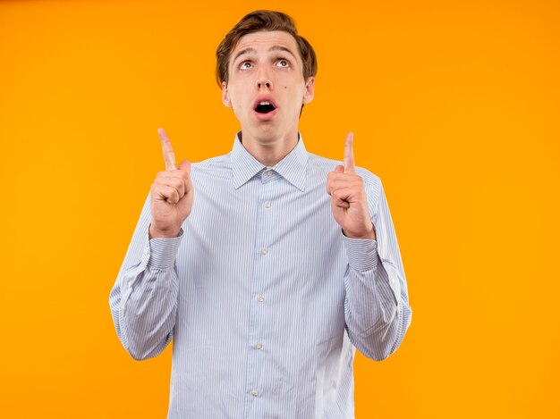 Young man in white shirt looking up pointing with index fingers up looking amazed and surprised standing over orange background