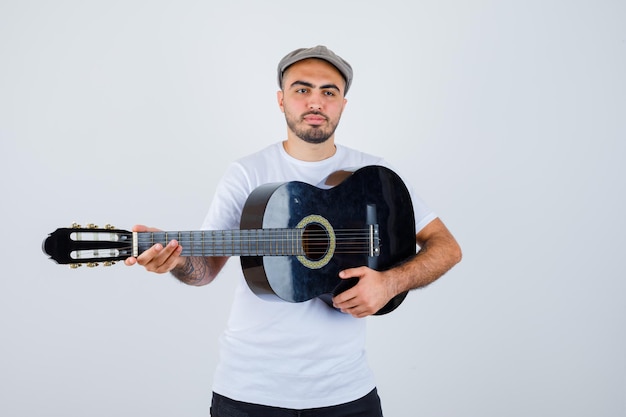 Young man in white t-shirt, black pants, grey cap playing guitar and looking serious