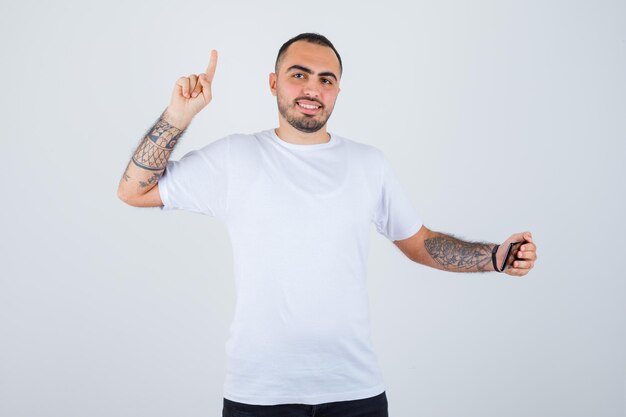 Young man in white t-shirt and black pants looking at something on phone and raising index finger in eureka gesture and looking happy