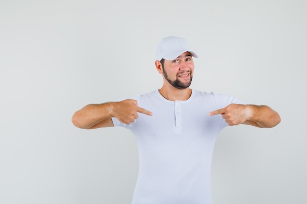 Free Photo young man in white t-shirt,cap pointing at himself and looking jolly , front view.