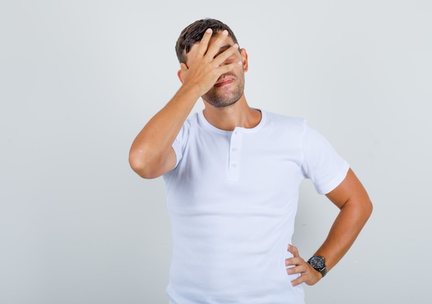 Young man in white t-shirt covering eyes with hand on waist and looking positive, front view.