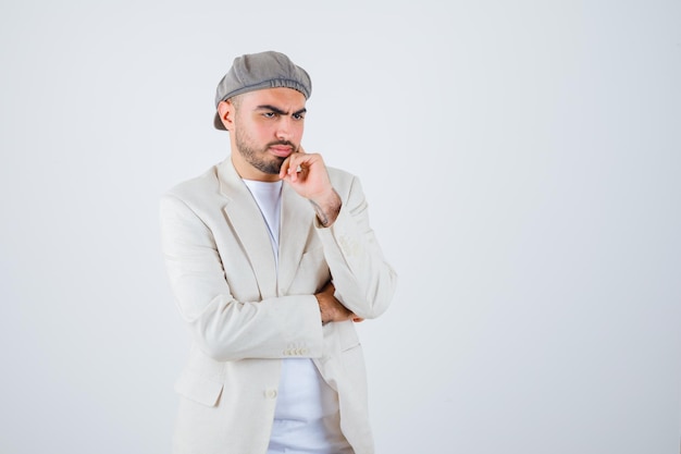 Young man in white t-shirt, jacket and gray cap standing in thinking pose and looking pensive