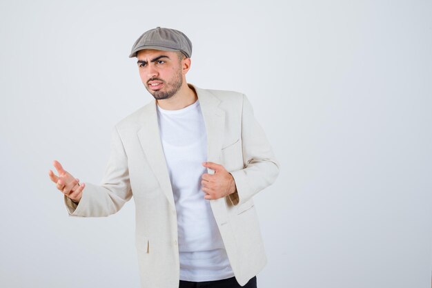 Young man in white t-shirt, jacket and gray cap stretching one hand as holding something and looking serious