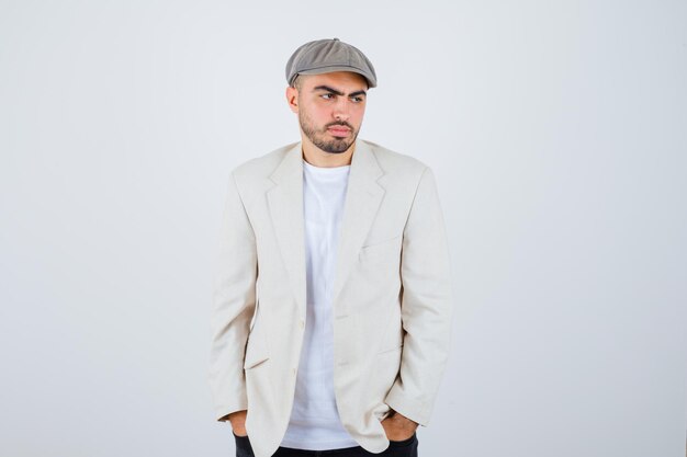 Young man in white t-shirt, jacket and grey cap holding hands in pocket and looking pensive