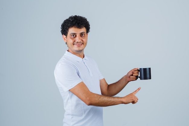 Young man in white t-shirt and jeans holding cup of tea while pointing to it and looking happy , front view.