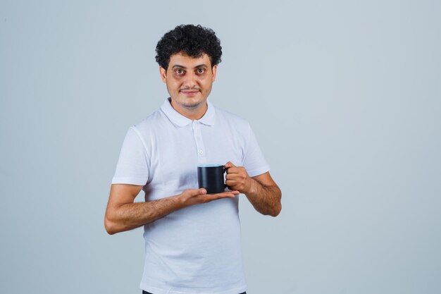 Young man in white t-shirt and jeans holding cup of tea with both hands and looking happy , front view.