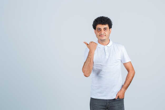 Young man in white t-shirt and jeans putting hand in pocket while pointing away and looking serious  , front view.