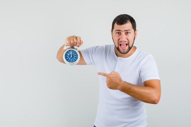 Young man in white t-shirt pointing at clock and looking impatient , front view.