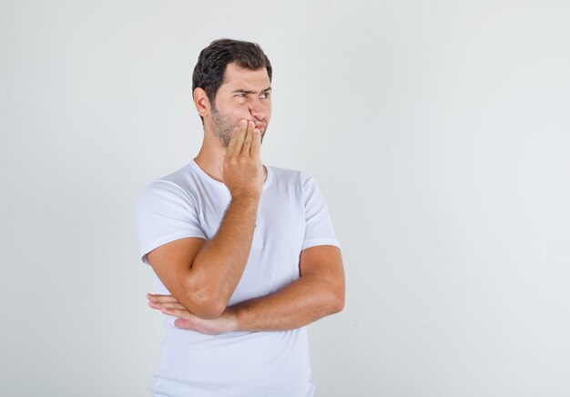 Young man in white t-shirt standing with hand near mouth and looking thoughtful