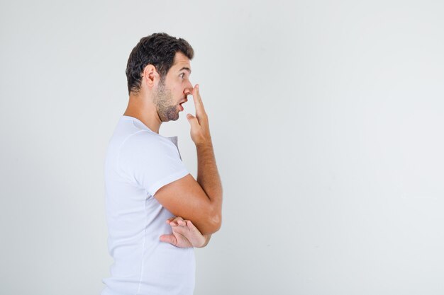 Young man in white t-shirt touching nose with hand and looking surprised .