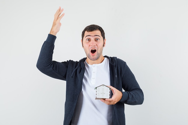 Young man in white t-shirt and zip-front black hoodie holding house model and raising hand in eureka gesture and looking excited , front view.