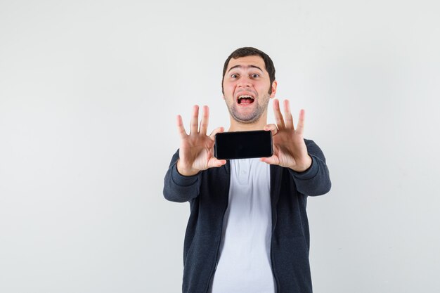 Young man in white t-shirt and zip-front black hoodie holding smartphone with both hands and looking happy , front view.