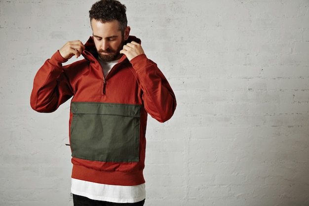 Free photo young man with beard putting on the hood of his stylish red and grey anorack isolated on white