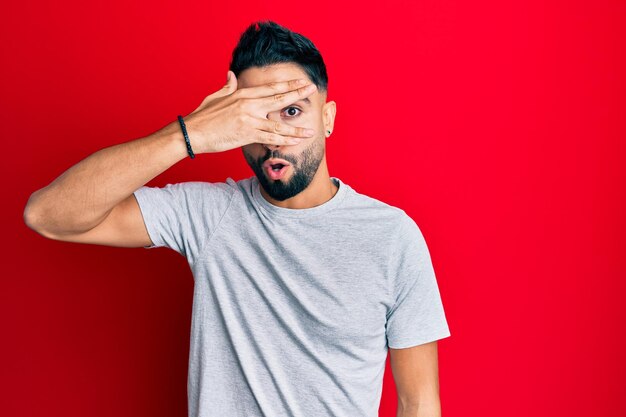 Young man with beard wearing casual white tshirt peeking in shock covering face and eyes with hand, looking through fingers afraid