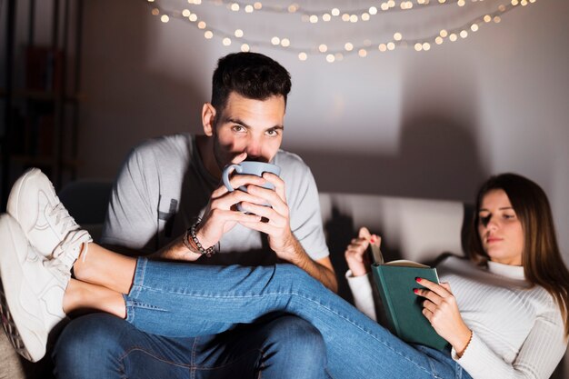 Young man with cup watching TV and woman with book on sofa