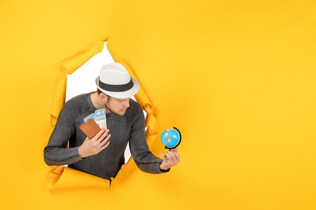 Free Photo young man with a hat holding foreign passport with ticket and small globe in a torn on yellow wall