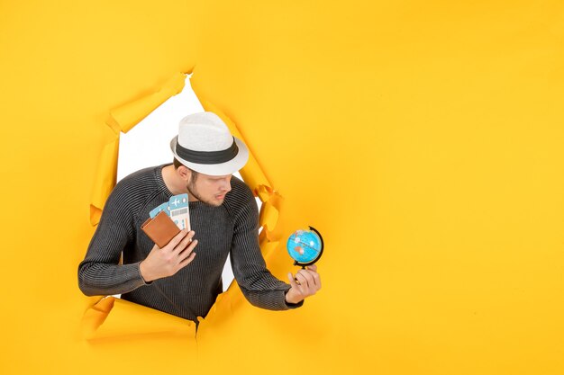 Young man with a hat holding foreign passport with ticket and small globe in a torn on yellow wall