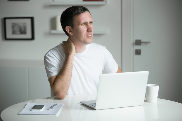 Free photo young man with his hands at his neck, sitting near desk