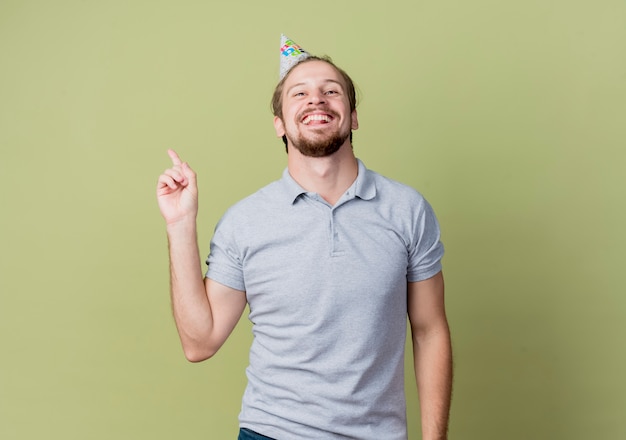 Young man with holiday cap celebrating birthday party happy and excited showing index finger over light wall