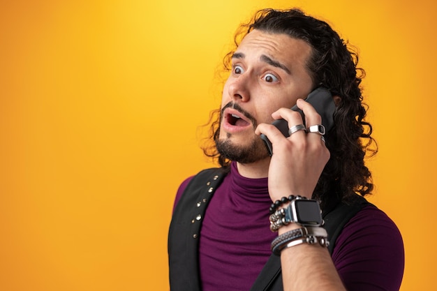 Free photo young man with long curly hair emotionally speaks on mobile phone against yellow background