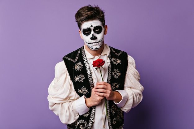 Free photo young man with sad look tenderly holds red flower at his chest. closeup portrait of brunet in halloween outfit.