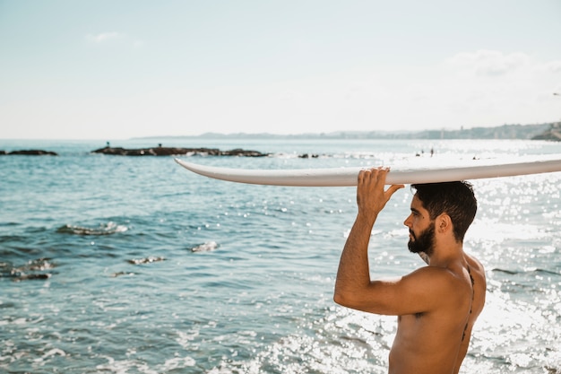 Young man with surf board on head on shore near water 