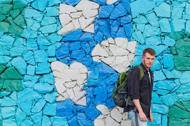 Young man with travel backpack standing near painted stone wall