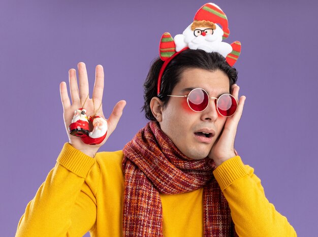 Young man with warm scarf around neck in yellow turtleneck and glasses with funny rim on head holding christmas toys  worried standing over purple wall