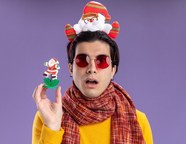 Young man with warm scarf around neck in yellow turtleneck with funny rim on head holding christmas toy  surprised standing over purple wall