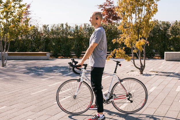 Free photo young man with wireless bluetooth standing with white bicycle in the park