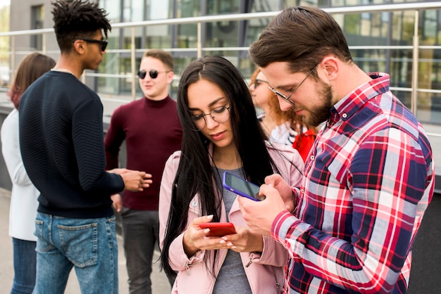 Young man and woman using smartphone with their friends standing behind outdoors