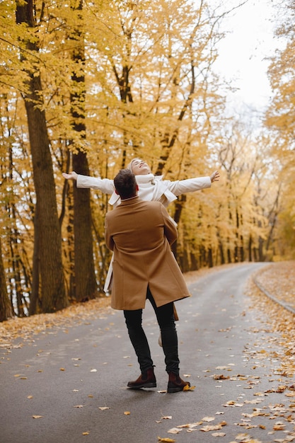 Free photo young man and woman walking outside wearing beige coats. blond woman and brunette man in autumn forest. man holding his wife in hands.