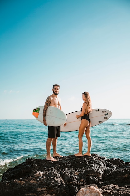 Young man and woman with surf boards on stone near water