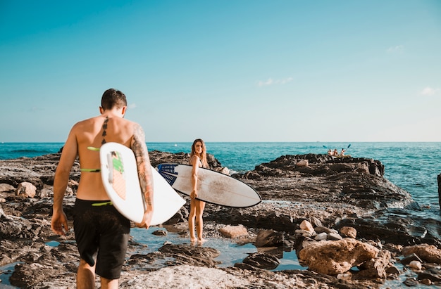 Young man and woman with surf boards on stone shore near water