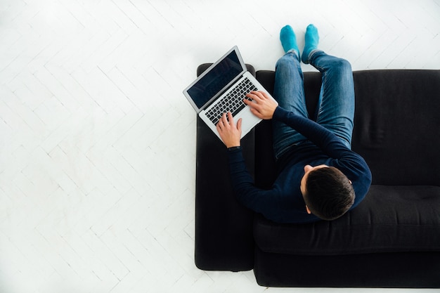 Young man working on laptop, sitting on black sofa, white floor. 