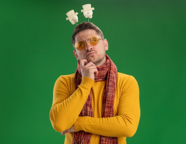 Young man in yellow turtleneck and glasses with warm scarf and funny rim on head looking up with pensive expression on face thinking standing over green wall