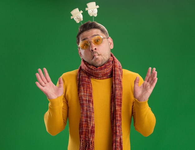 Young man in yellow turtleneck and glasses with warm scarf and funny rim on head standing over green wall