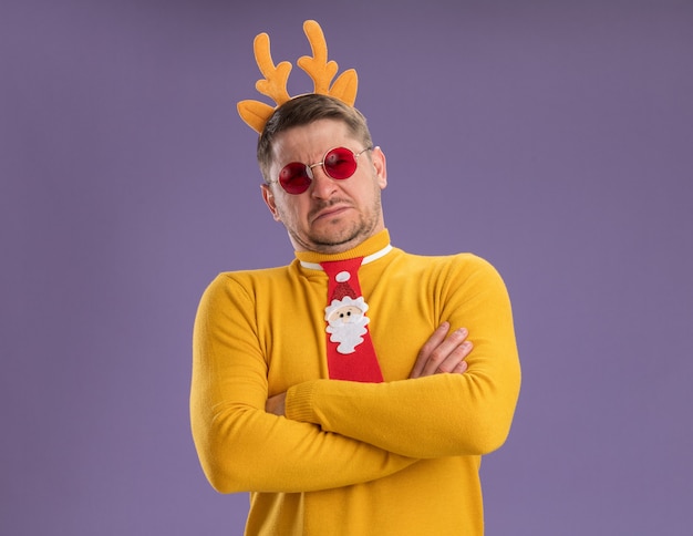 Free Photo young man in yellow turtleneck and red glasses wearing funny red tie and rim with deer horns on head looking at camera displeased with arms crossed standing over purple background