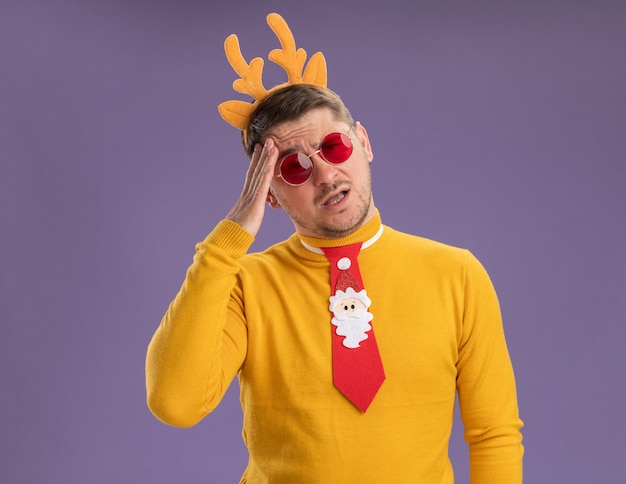 Free Photo young man in yellow turtleneck and red glasses wearing funny red tie and rim with deer horns on head looking confused and very anxious standing over purple background