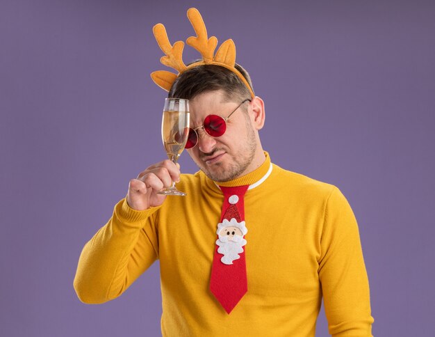 Young man in yellow turtleneck and red glasses wearing funny red tie and rim with deer horns holding glass of champagne looking confused and very disappointed standing over purple background