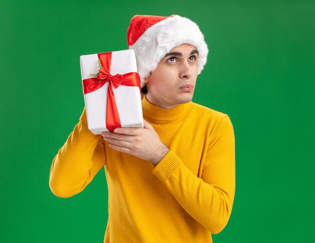 Young man in yellow turtleneck and santa hat holding a present over his ear looking intrigued standing over green background