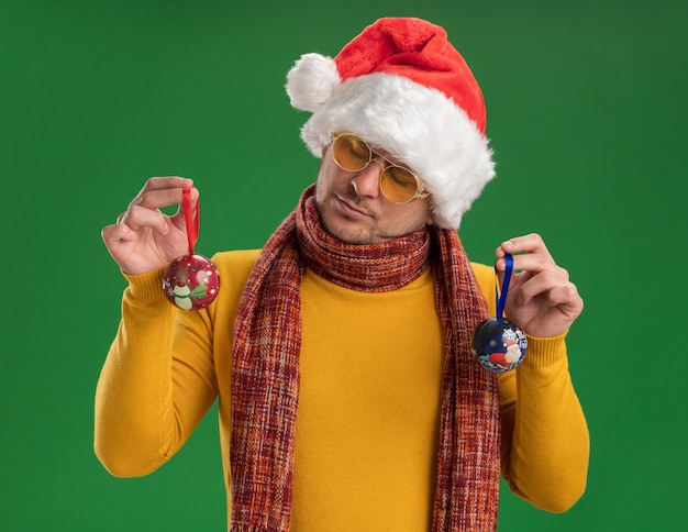 Free Photo young man in yellow turtleneck and santa hat wearing glasses holding toys for christmas tree looking at them with serious face standing over green wall