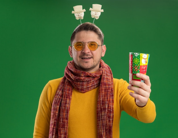 Free photo young man in yellow turtleneck with warm scarf and glasses wearing funny rim on head holding colorful cup  with smile on face standing over green wall